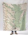 A person holding a blanket that features a vintage map design of the Virginia side of the Chesapeake Bay area with various town names marked in green on a beige background.